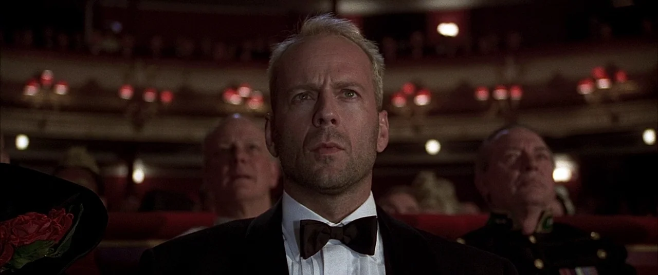 The Fifth Element (1997) movie still: medium — A dimly lit theater interior with red velvet seats and blurred balcony seating background. A man in a…