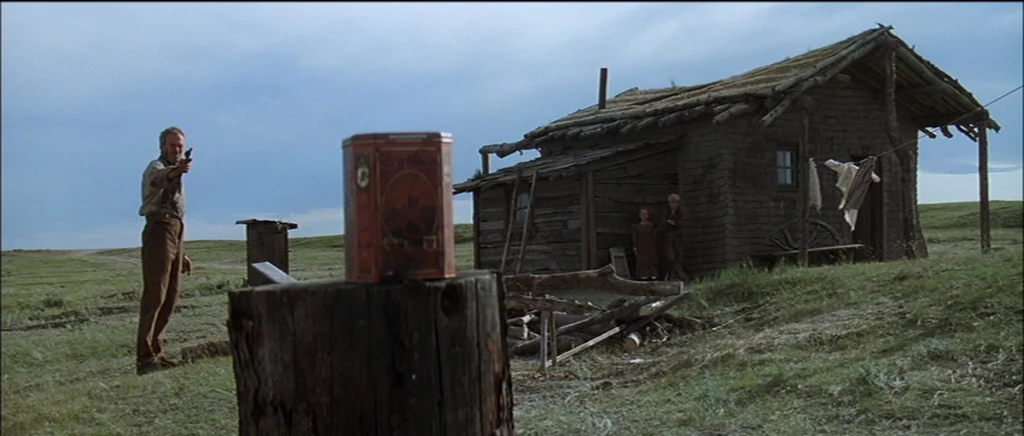 Unforgiven (1992) movie still: wide — A remote prairie homestead features a weathered wooden shack under a gray overcast sky. A man stands in the…