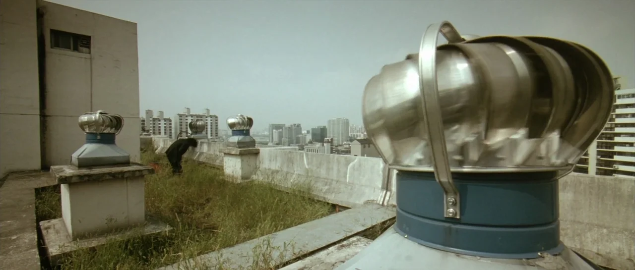 Oldboy (2003) movie still: wide — A rooftop covered in overgrown grass features several metallic rotating turbine vents. A lone person in a dark…