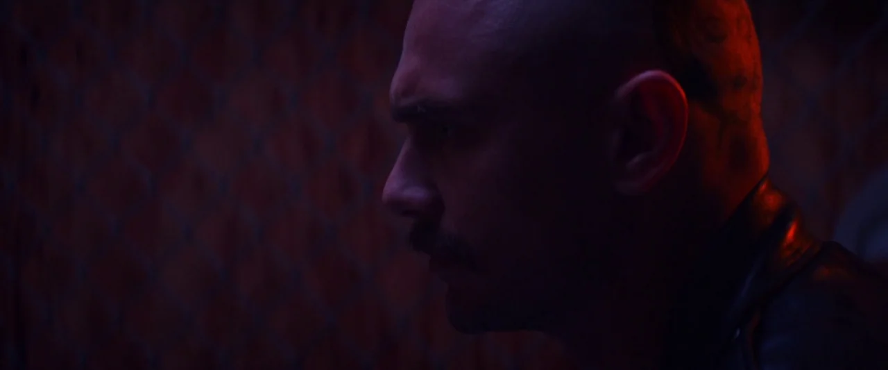 Zeroville (2019) movie still: closeup — A dark space features a chain-link fence backdrop under colored light. A bald man with a mustache sits in…