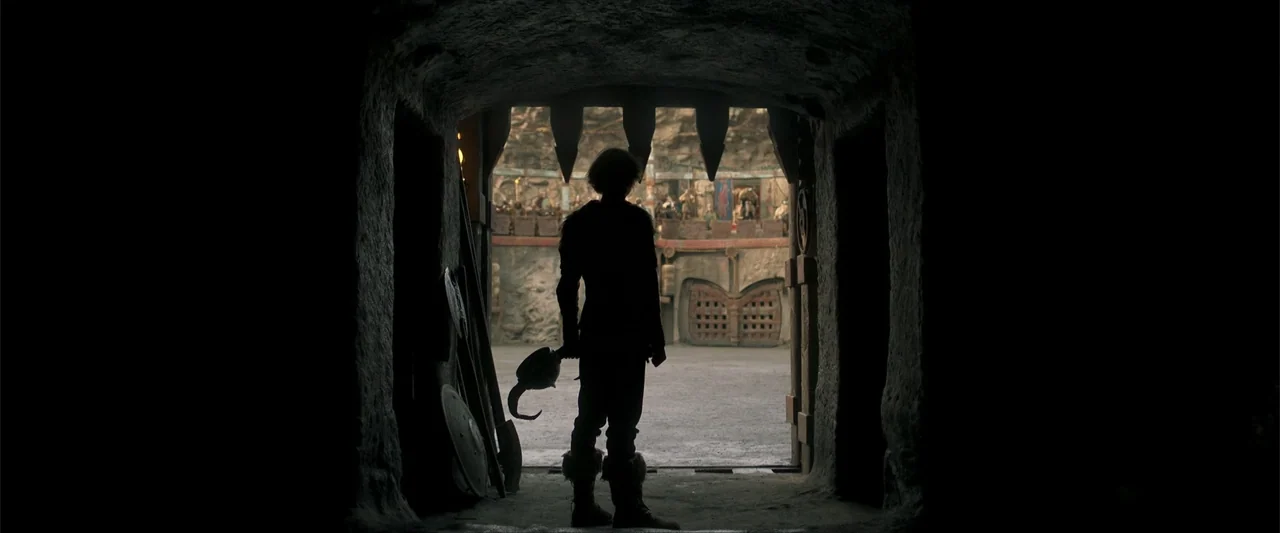 How to Train Your Dragon (2025) movie still: fullbody — A dark stone tunnel frames an view of an sunlit outdoor circular arena. A youth stands in…