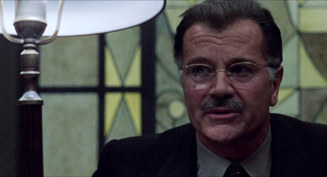 The Night Porter (1974) movie still: closeup — An interior space features a leaded glass window backdrop with geometric patterns. A man in a suit…