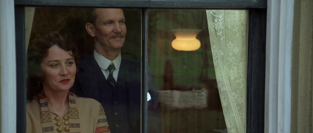 Seabiscuit (2003) movie still: medium — A man and a woman stand behind a window pane looking outwards at dusk. The interior lamp reflects faintly…