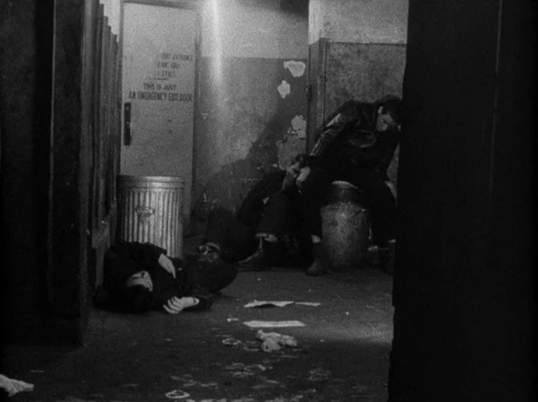 Shadows (1958) movie still: wide — A dimly lit concrete stairwell area features peeling walls and a metal trash bin. Three young men are slumped on…