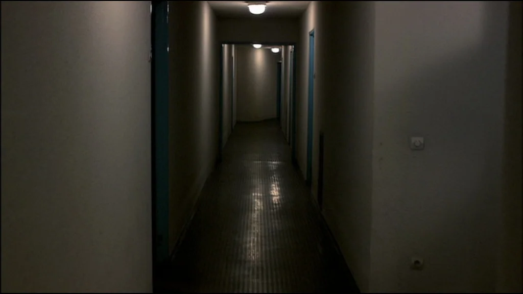 Caché (Hidden) (2005) movie still: establishing — A long, dimly lit residential hallway features plain white walls and a reflective floor. Multiple…