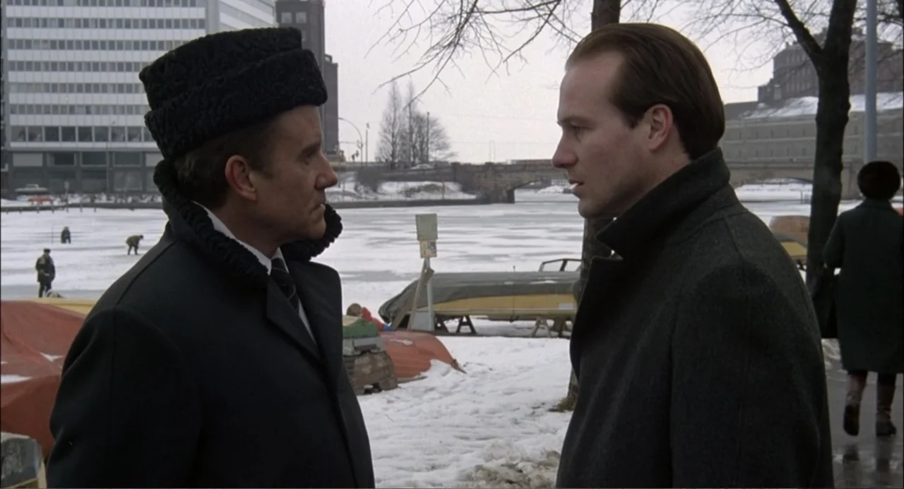 Gorky Park (1983) movie still: medium — A snowy, frozen waterfront area features distant buildings and leafless trees on an overcast day. Two men in…