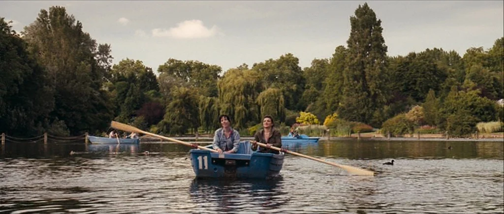 Happy-Go-Lucky (2008) movie still: wide — A calm lake features a blue rental boat numbered 11 with two women inside. The background shows lush green…