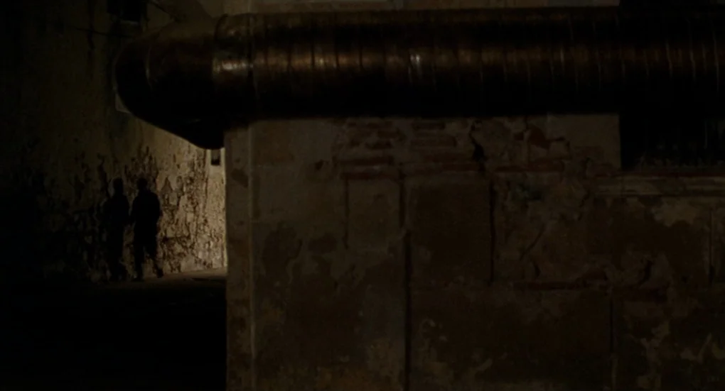 Furia (1999) movie still: wide — A dark narrow alleyway features an old crumbling brick wall and a metallic conduit pipe. Two silhouetted figures…