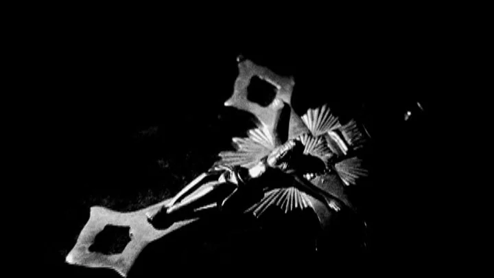 Dracula: Pages from a Virgin’s Diary (2002) movie still: closeup — A metal crucifix is suspended against a completely dark, featureless black…