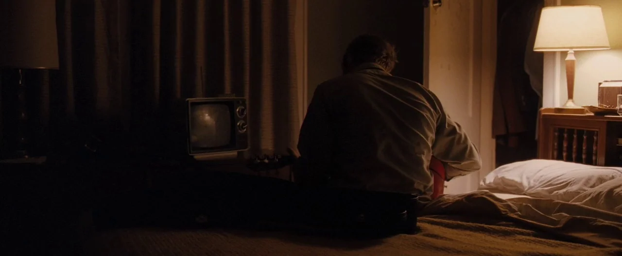 Ain’t Them Bodies Saints (2013) movie still: medium — A dimly lit motel room featuring a bed with rumpled white linens and dark curtains. A man sits…