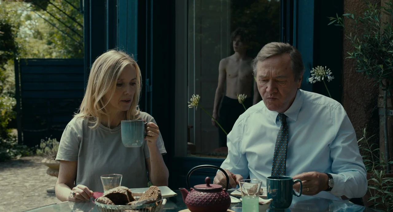 Last Summer (2023) movie still: medium — A man and woman sit at a glass table with breakfast items on a patio. A shirtless young man stands in the…