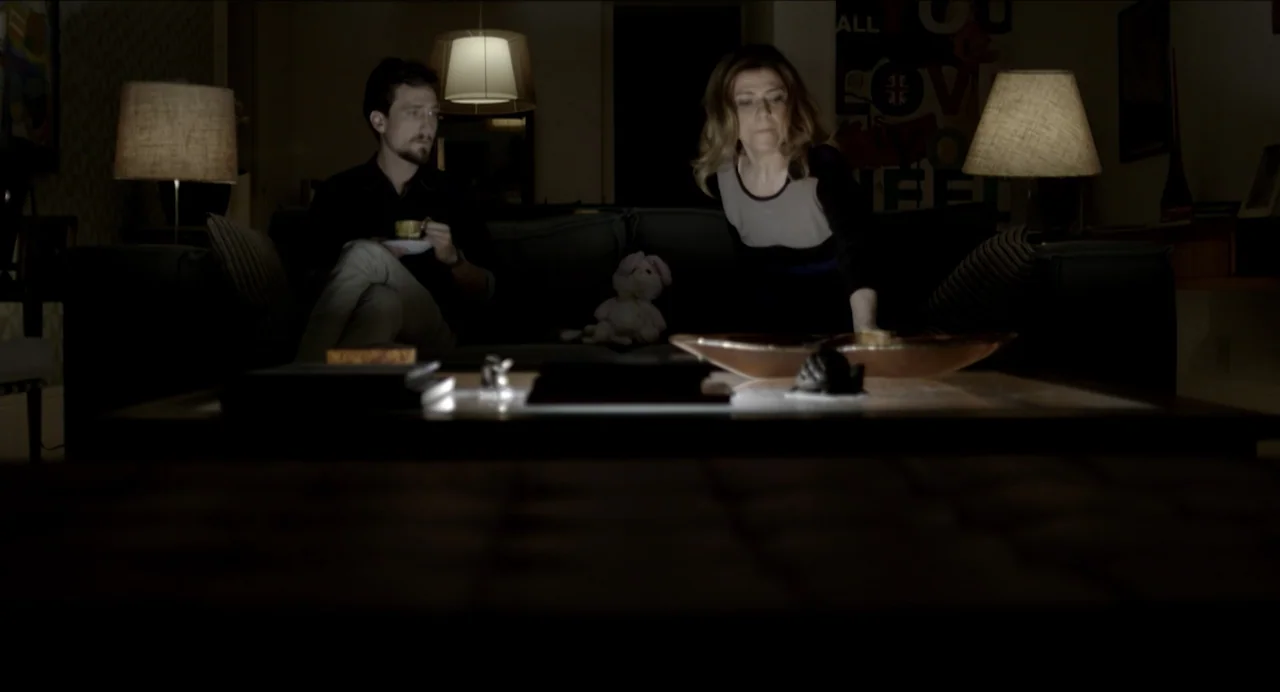 Suspension Points (2022) movie still: medium — A dimly lit living room with dark walls and two table lamps on either side. A man sits on a sofa with…