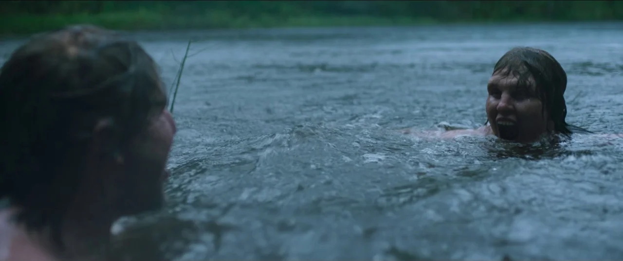 Border movie still: medium — Two individuals are waist-deep in a murky, dark-water river surrounded by distant green foliage. Both people face each…