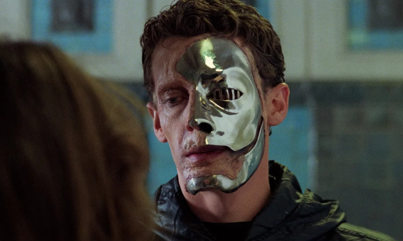 Demons (1985) movie still: closeup — A man in a dim corridor faces another person while wearing a reflective metal mask. One half of his face is…