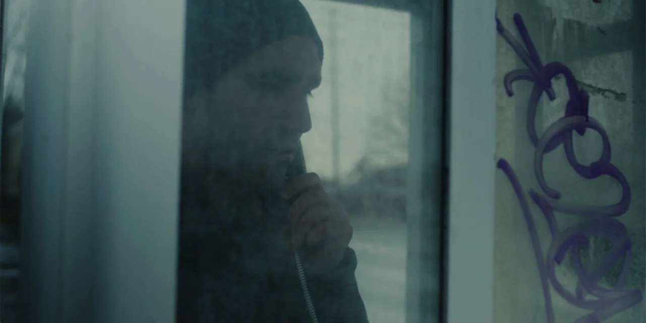 Lobos (2019) movie still: medium — A man stands inside a glass-walled phone booth marked with purple graffiti tags. He holds the telephone receiver…