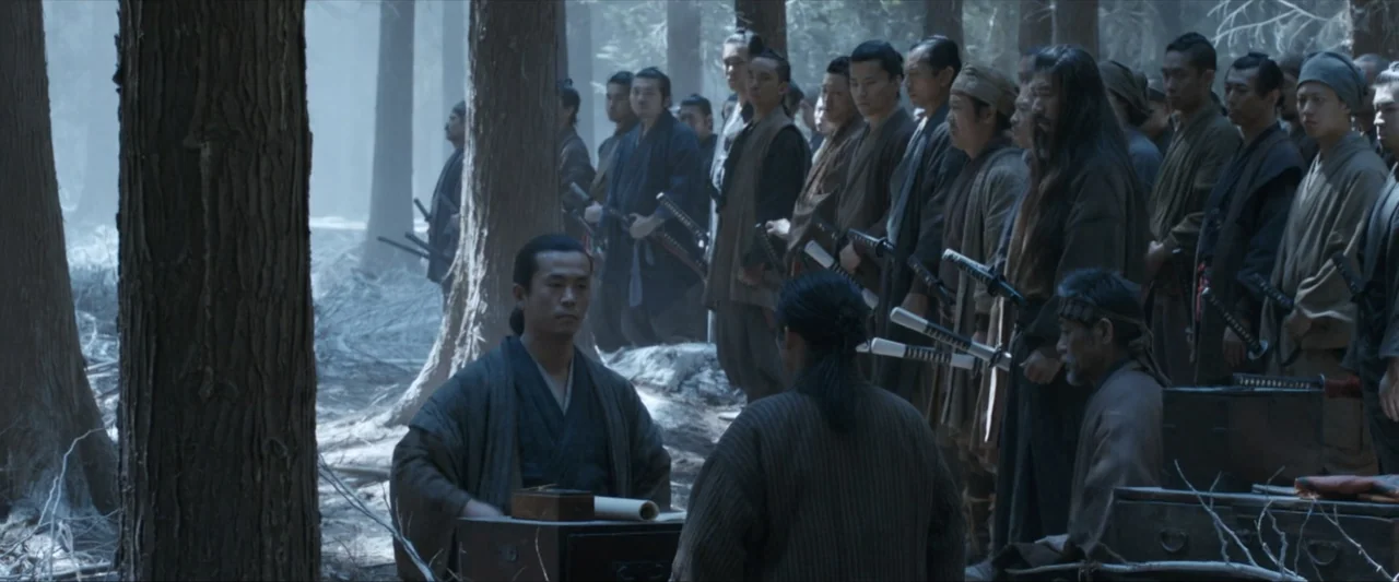 47 Ronin (2013) movie still: wide — A large group of armed men stands within a dense, hazy forest clearing. A central figure sits at a small wooden…