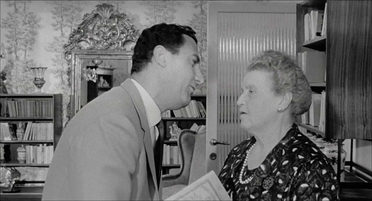 Il Boom (1963) movie still: medium — A residential library features floor-to-ceiling bookshelves and ornate floral wallpaper behind a mirror. A man…