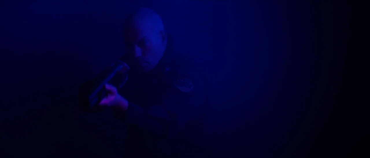 Dog Eat Dog (2016) movie still: medium — A dark outdoor space is filled with blue light and atmospheric haze. A police officer holds a firearm…