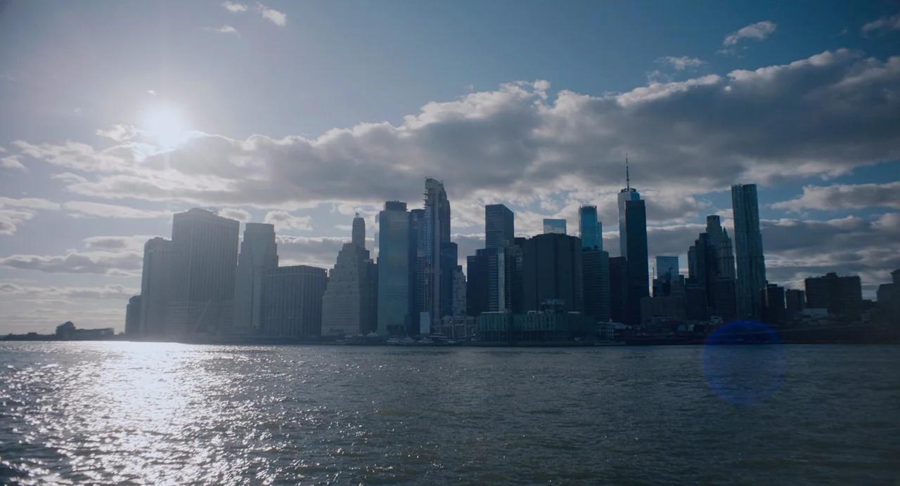 She Said (2022) movie still: establishing — A view of the lower Manhattan skyline from the water under a bright sun. The dense collection of…
