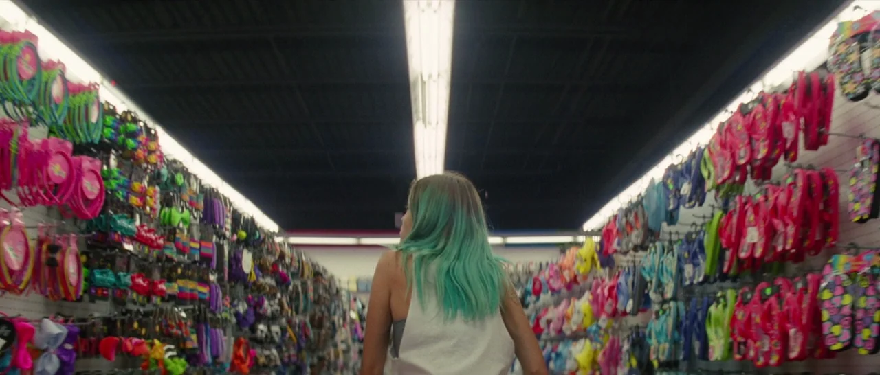 The Florida Project (2017) movie still: medium — A brightly lit retail aisle is lined with dense rows of colorful hanging merchandise. A young woman…