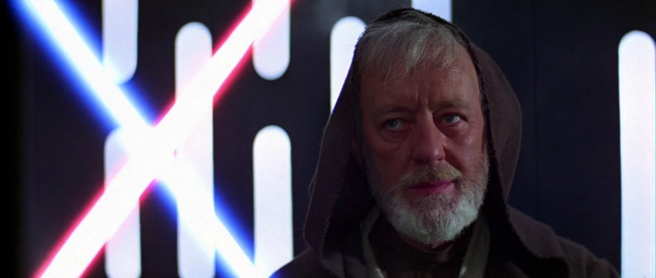 Star Wars (1977) movie still: closeup — A dark, industrial interior features glowing vertical light panels on a black wall. An elderly man with a…