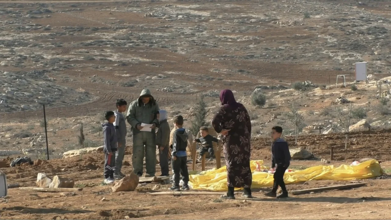 No Other Land (2024) movie still: wide — An arid, rocky hillside features a group of individuals standing on patches of dirt. An adult in a brown…
