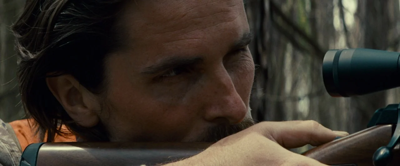 Out of the Furnace (2013) movie still: closeup — A man lies prone in a woodland area with dense, out-of-focus trees behind him. He rests his cheek…
