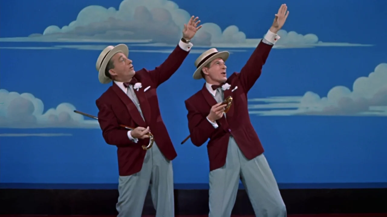 White Christmas (1954) movie still: fullbody — A theatrical stage with a painted backdrop of clouds against a bright blue sky. Two men in matching…