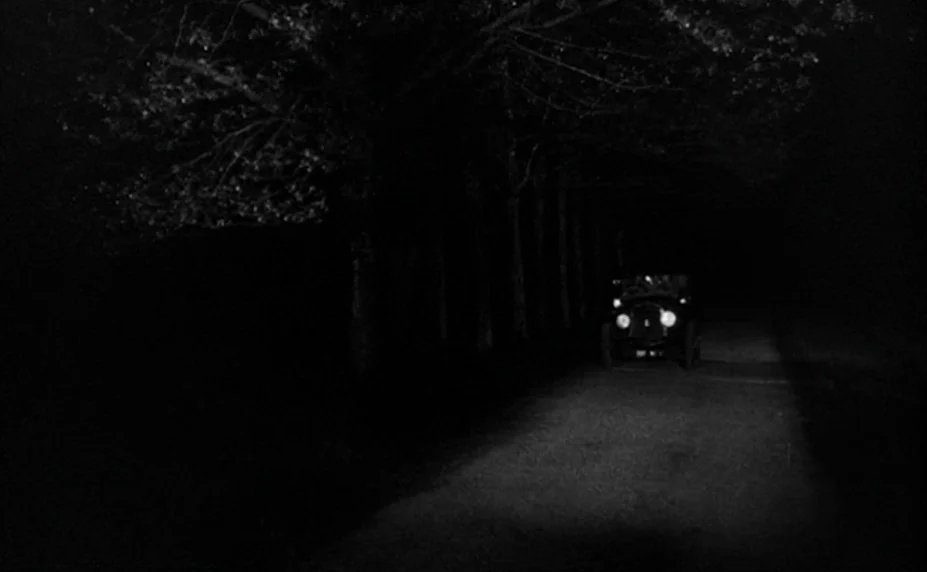 Judex (1966) movie still: wide — A dark country road is bordered by a dense line of trees at night. A lone vintage car approaches with its…