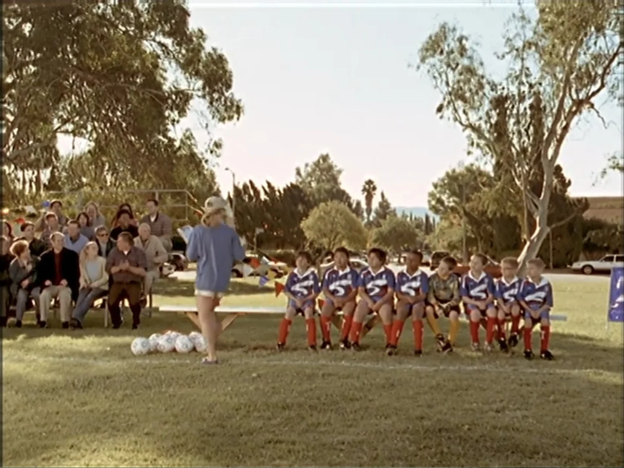 Just 4 Kicks (2003) movie still: wide — A sunny soccer field features a row of children sitting on a bench. A person stands in front of them while…