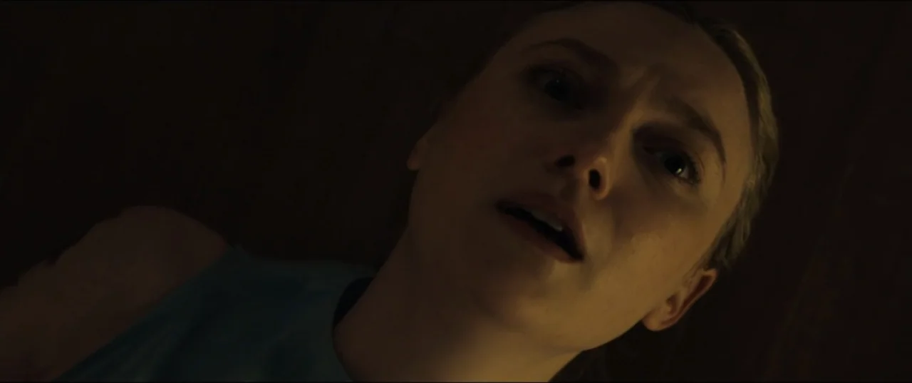 Vicious (2025) movie still: closeup — A dimly lit interior room with dark, obscured surroundings. A young woman lying down looks up toward the…
