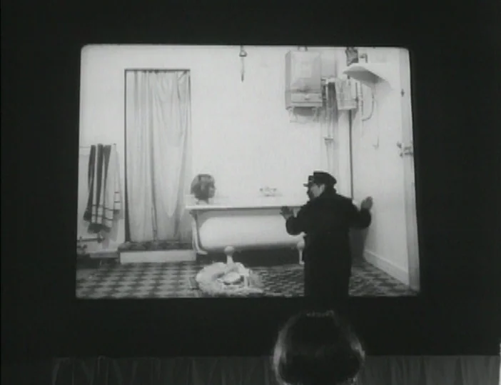 Les Carabiniers (1963) movie still: medium — A spartan bathroom with checkerboard flooring and a white pedestal bathtub against the wall. A man in a…