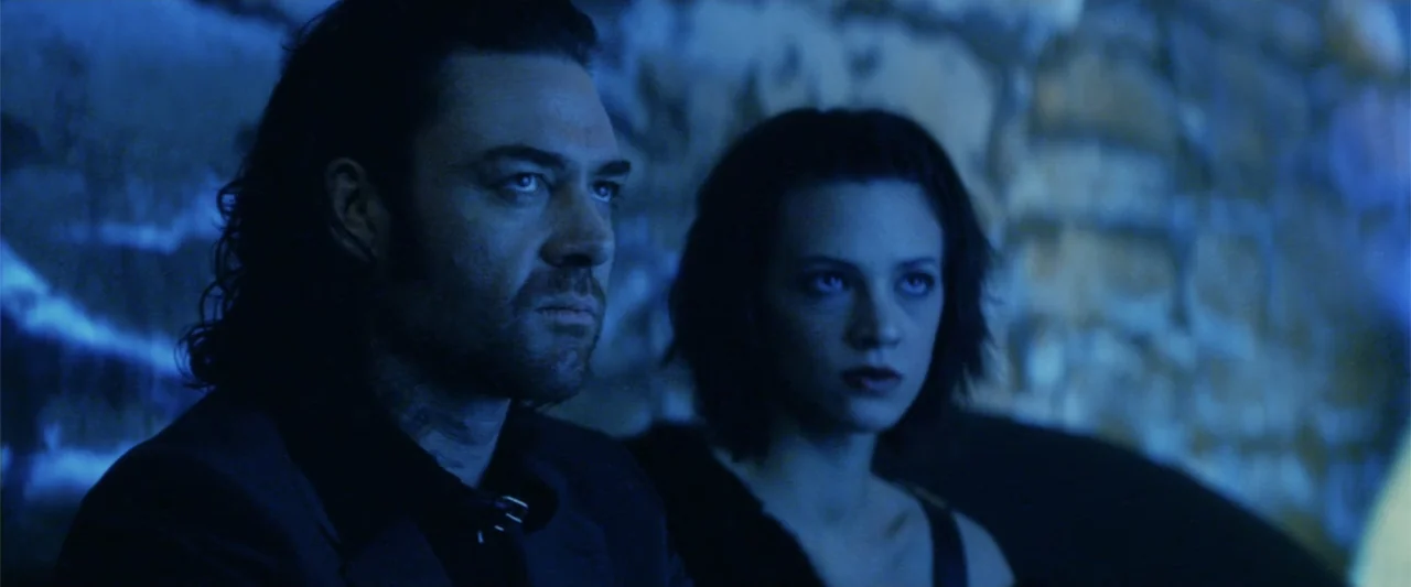xXx (2002) movie still: medium — A dark indoor space with a textured wall illuminated by cool blue light. A man and a woman are seated together…