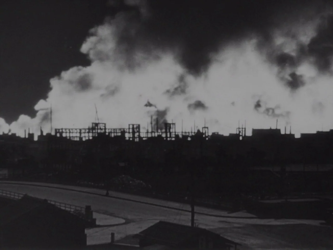 Godzilla Raids Again (1955) movie still: wide — A dark industrial skyline is silhouetted against a massive, illuminated cloud of smoke at night. The…