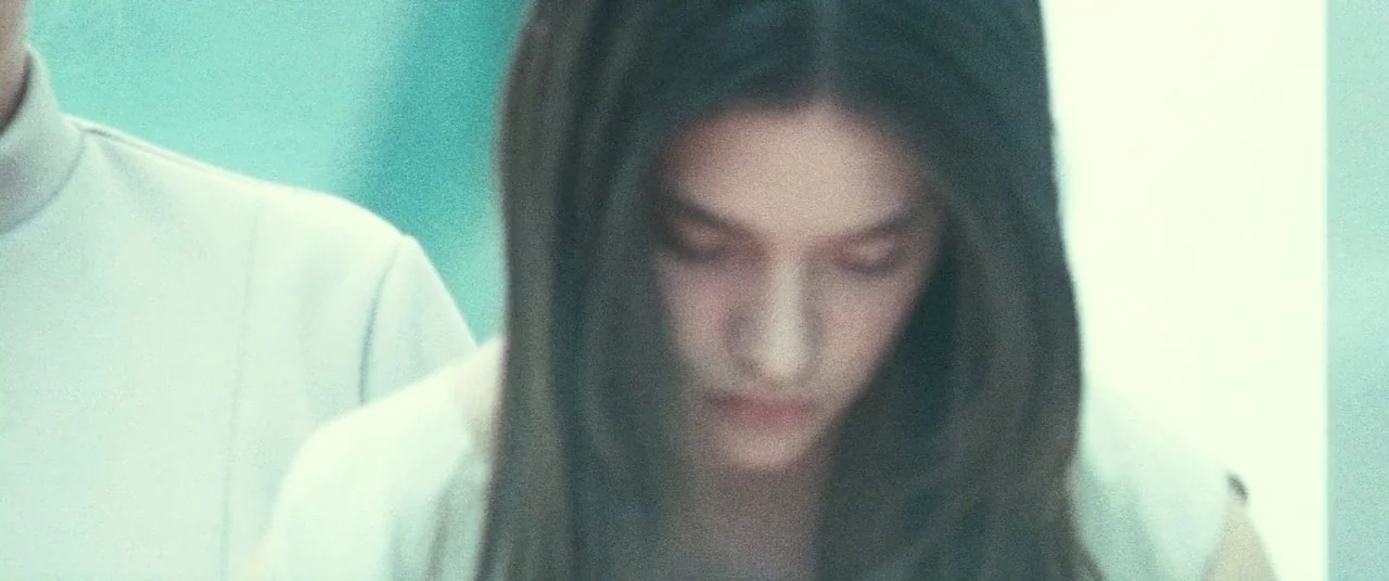 Beyond The Black Rainbow (2010) movie still: closeup — A sterile interior environment with bright white walls and cool blue lighting tones. A young…