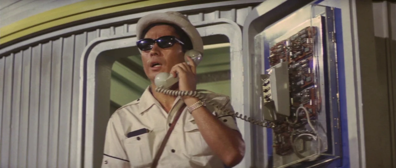 Son of Godzilla (1967) movie still: medium — An outdoor phone station with a transparent casing revealing internal electronic circuit boards. A man…
