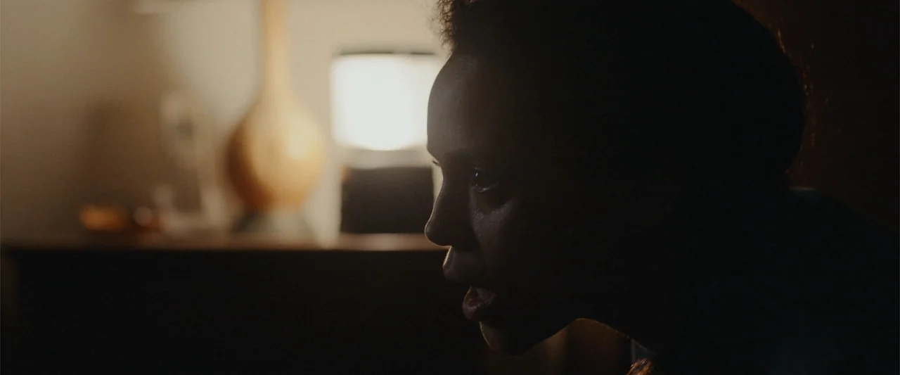 It Comes at Night (2017) movie still: closeup — A dimly lit interior space features a glowing lamp in the background. A woman looks off-screen with…