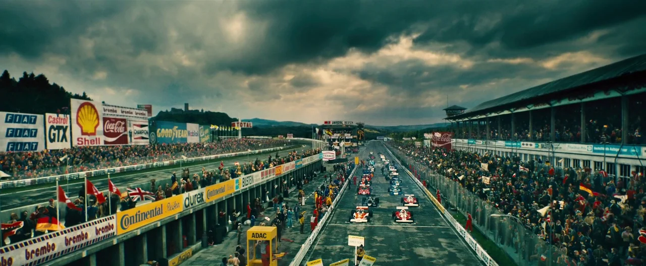 Rush (2013) movie still: wide — A high-angle view of a large auto racing circuit under a dark, overcast sky. Numerous classic Formula One cars sit…