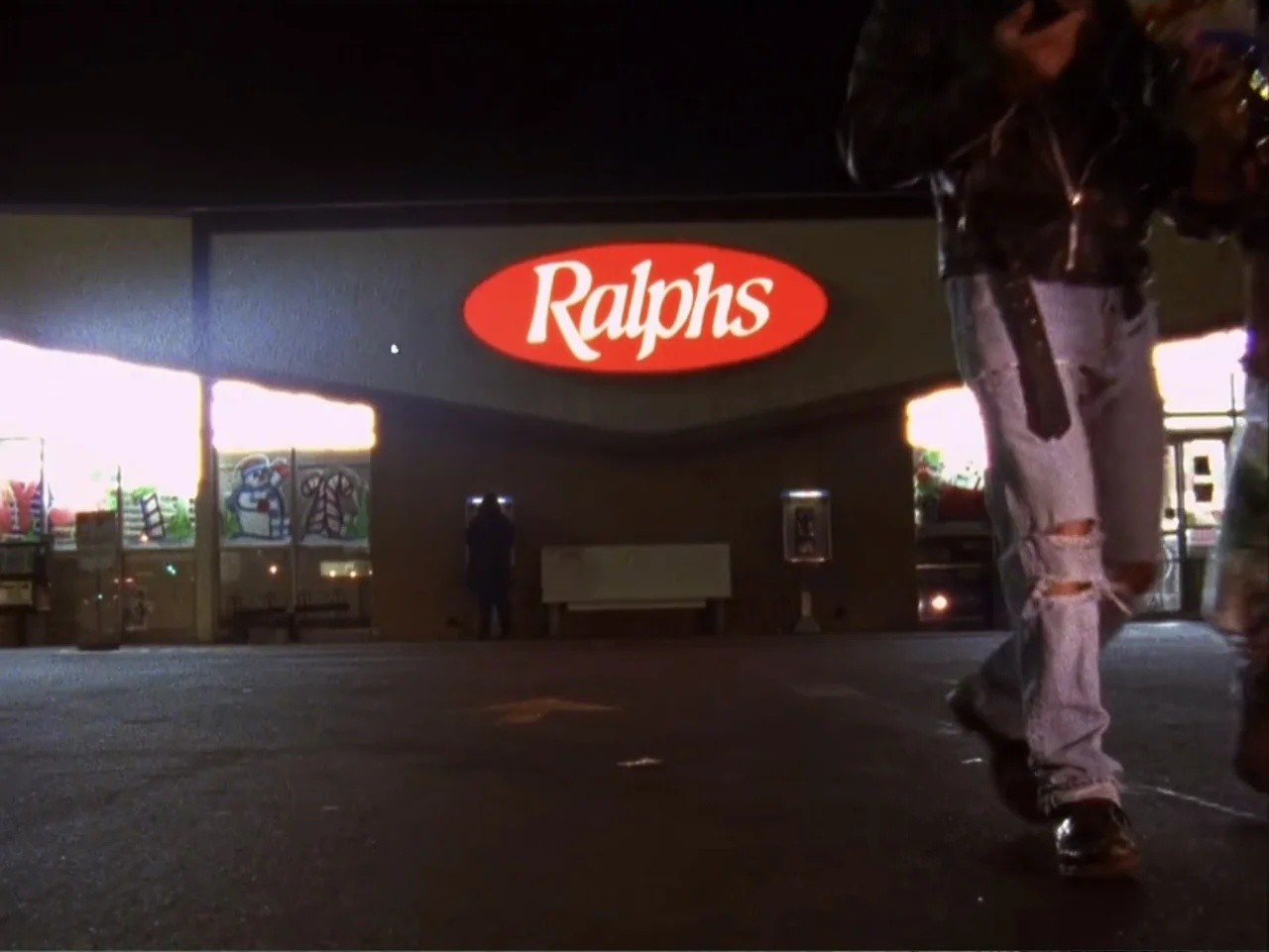 The Living End (1992) movie still: medium — A dark, paved grocery store parking lot at night with a prominent glowing sign. A man in a leather…