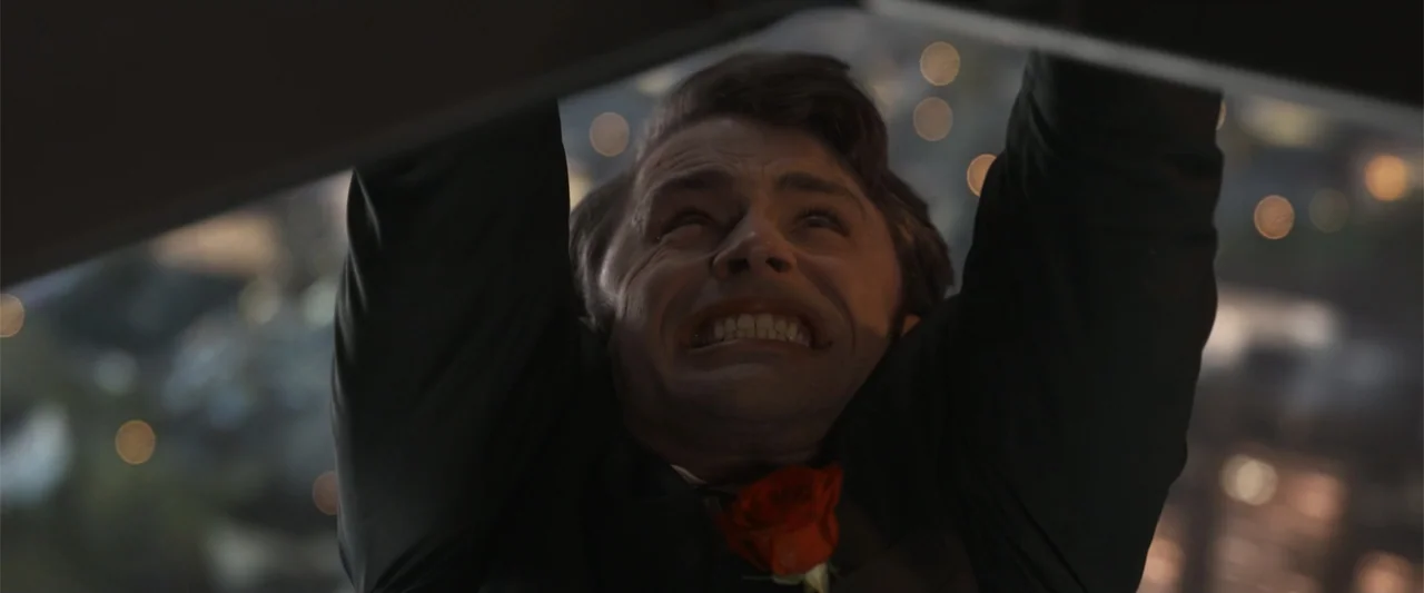 Final Destination Bloodlines (2025) movie still: closeup — A man hangs from a ledge high above a city at night. The urban background is out of focus…
