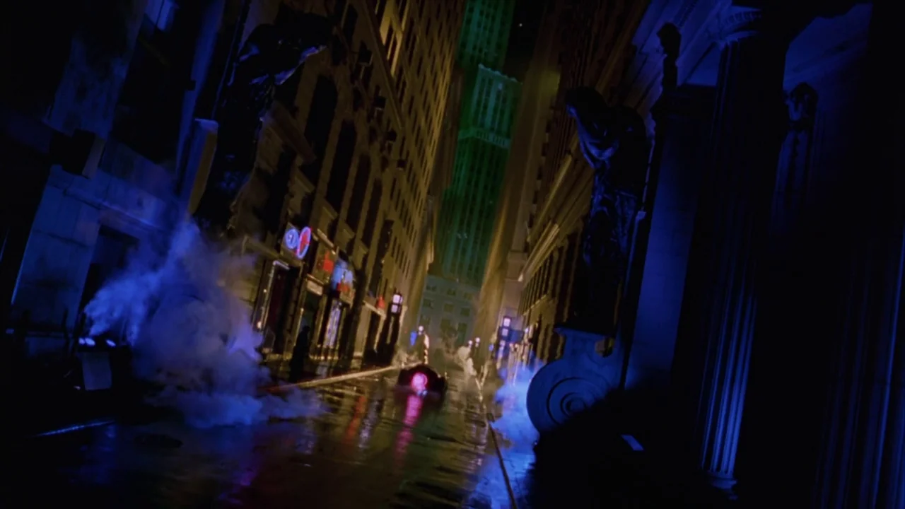 Batman Forever (1995) movie still: establishing — A dark, narrow urban canyon is flanked by towering buildings with stone carvings. Steam rises from…