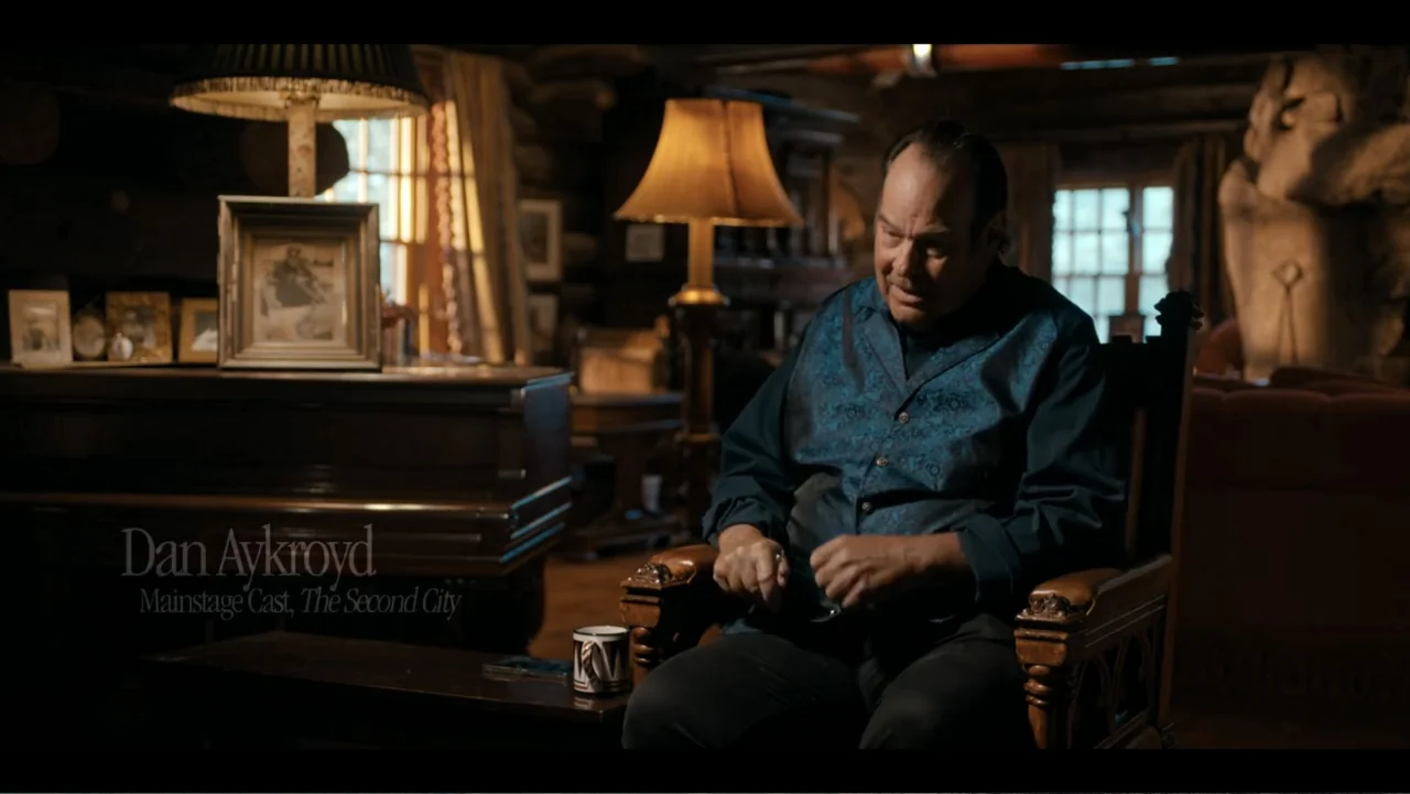 John Candy: I Like Me (2025) movie still: medium — A dimly lit log cabin space contains antique wood furniture and framed personal photos. A…