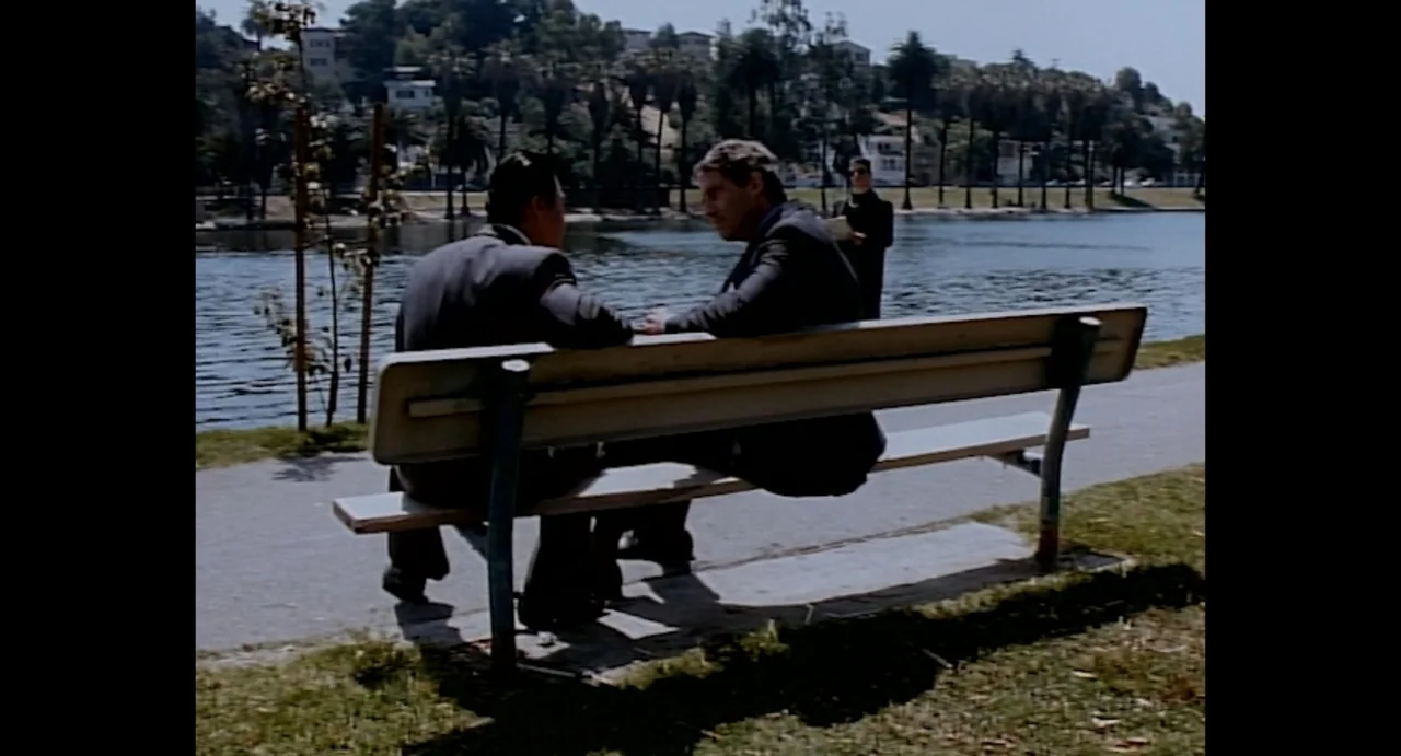 American Yakuza (1993) movie still: medium — A public park path by a lake with a wooden bench in the foreground. Two men in business suits sit…