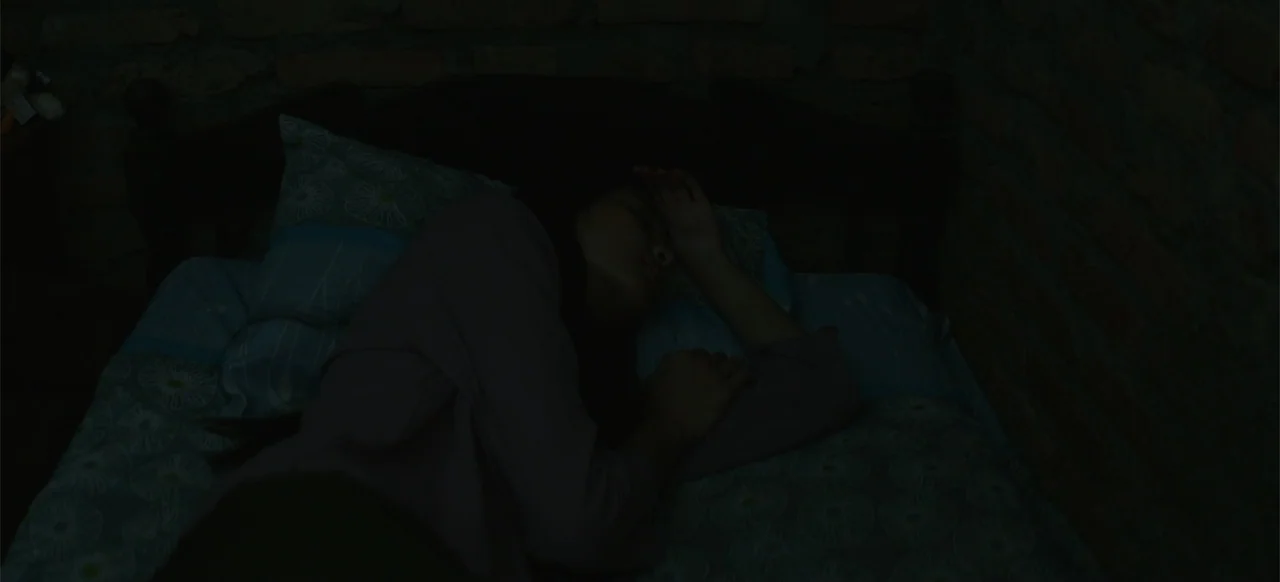 A Thousand Days (2023) movie still: medium — A dimly lit room features a person sleeping on a bed against a brick wall. The subject lies on their…