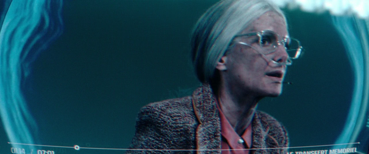 Oxygen (2021) movie still: medium — An older woman with silver hair is captured within a blue-tinted digital video feed. The frame displays playback…
