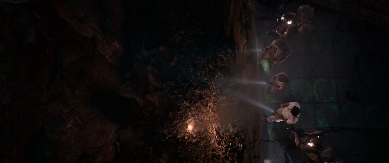 Tomb Raider (2018) movie still: wide — A group of five people stand on a stone ledge overlooking a deep dark pit. They point flashlights downward,…