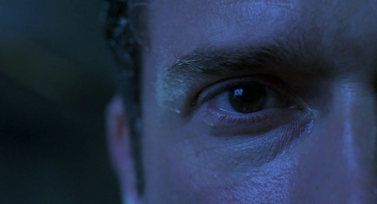 Resident Evil (2002) movie still: closeup — A close-up view focuses on a human eye in a darkened environment. Blue-toned light reflects off the iris…