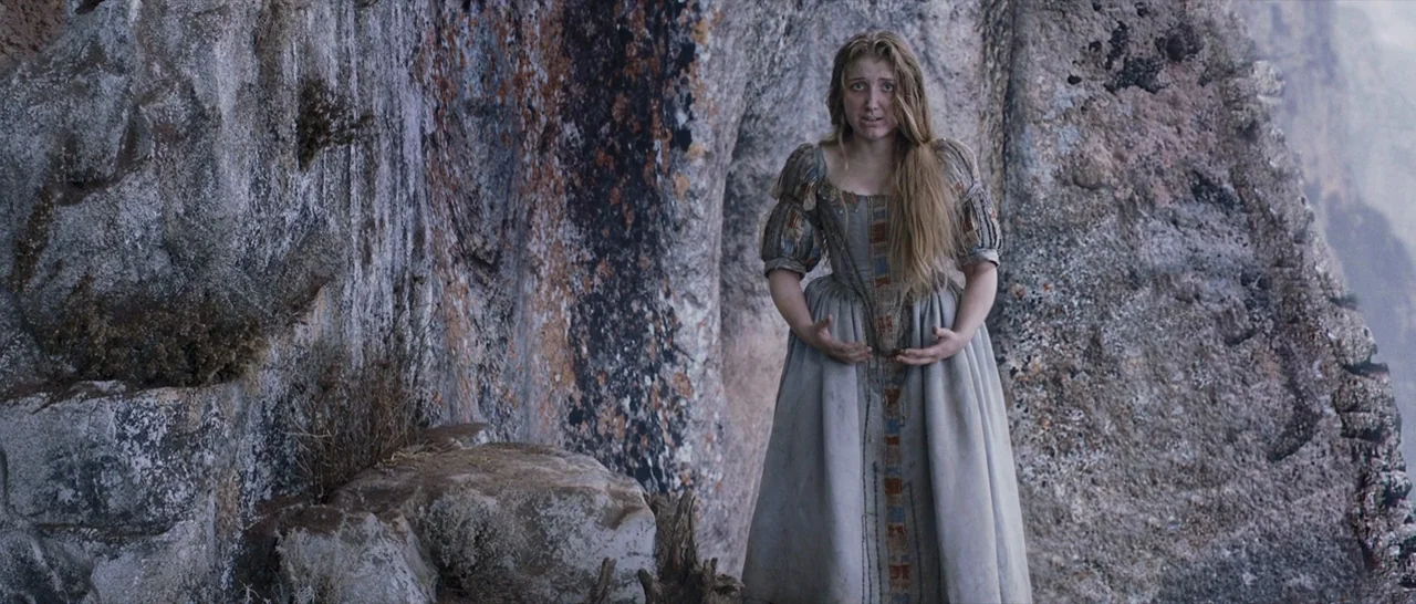 Tale of Tales (2015) movie still: fullbody — A woman stands before a textured, light-grey rock wall on a steep cliff face. The woman wears a…