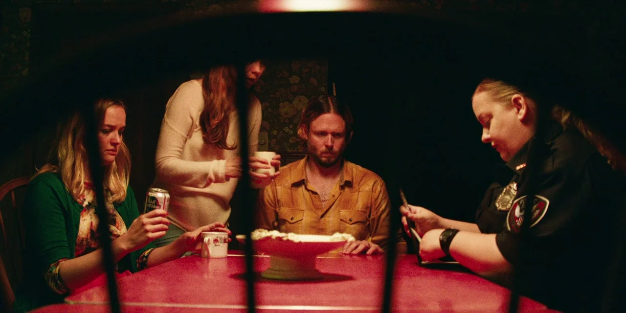 The Death of Dick Long (2019) movie still: medium — A dark dining room with patterned floral wallpaper features a bright red table. A police officer…