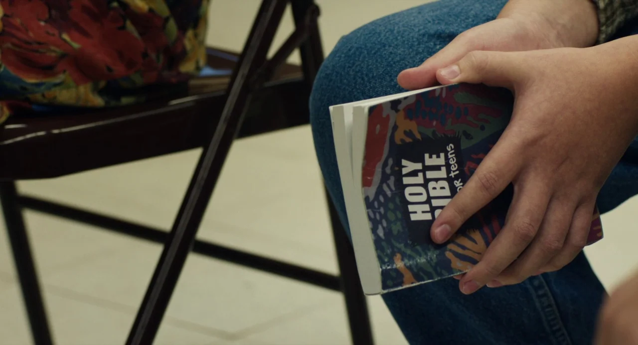 The Miseducation of Cameron Post (2018) movie still: closeup — A close-up view shows a person sitting on a metal folding chair. They hold a small…